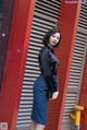 A woman in a black shirt and blue skirt leaning against a red wall.