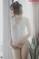 A woman in a white tank top and panties leaning against a window.