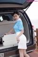 A woman in a blue shirt and white skirt standing next to a car.