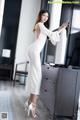 A woman in a white dress leaning against a dresser.