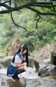 A woman in a school uniform crouching on a rock by a stream.