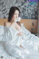 A woman in a white robe sitting on a bed drinking from a cup.