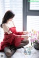 A woman in a red dress sitting on a bed next to a window.