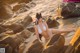 A woman in a white bikini sitting on some rocks by the water.