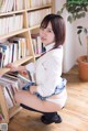 A woman in a school uniform is sitting on the floor in front of a bookshelf.